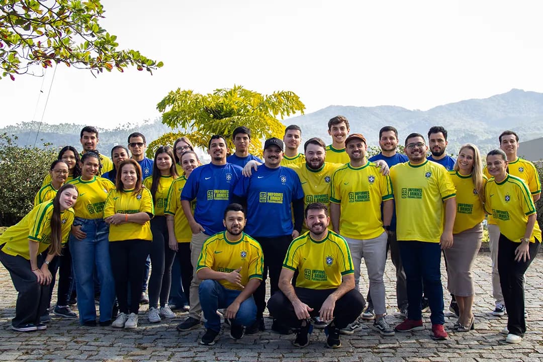 Equipe da T&M reunida ao ar livre, vestindo camisetas amarelas e azuis da empresa, com as montanhas de Brusque ao fundo